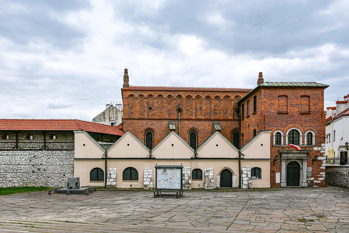 The Old Synagogue in Kazimierz, a red-brick building with a tiled roof and a row of arched windows, seen across a paved square.